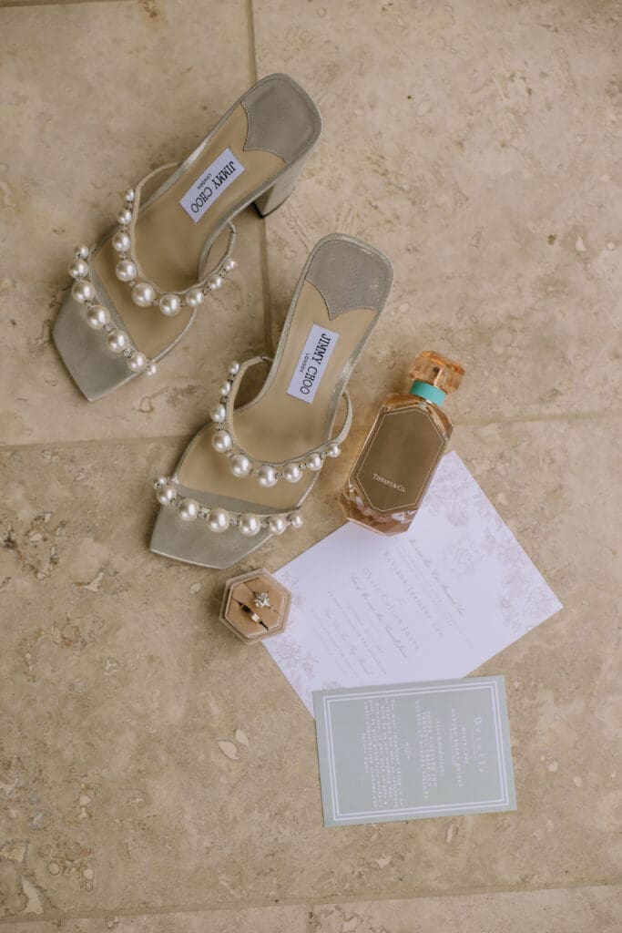 Wedding flat lay of: Jimmy Choo shoes, perfume, wedding invites, and rings.