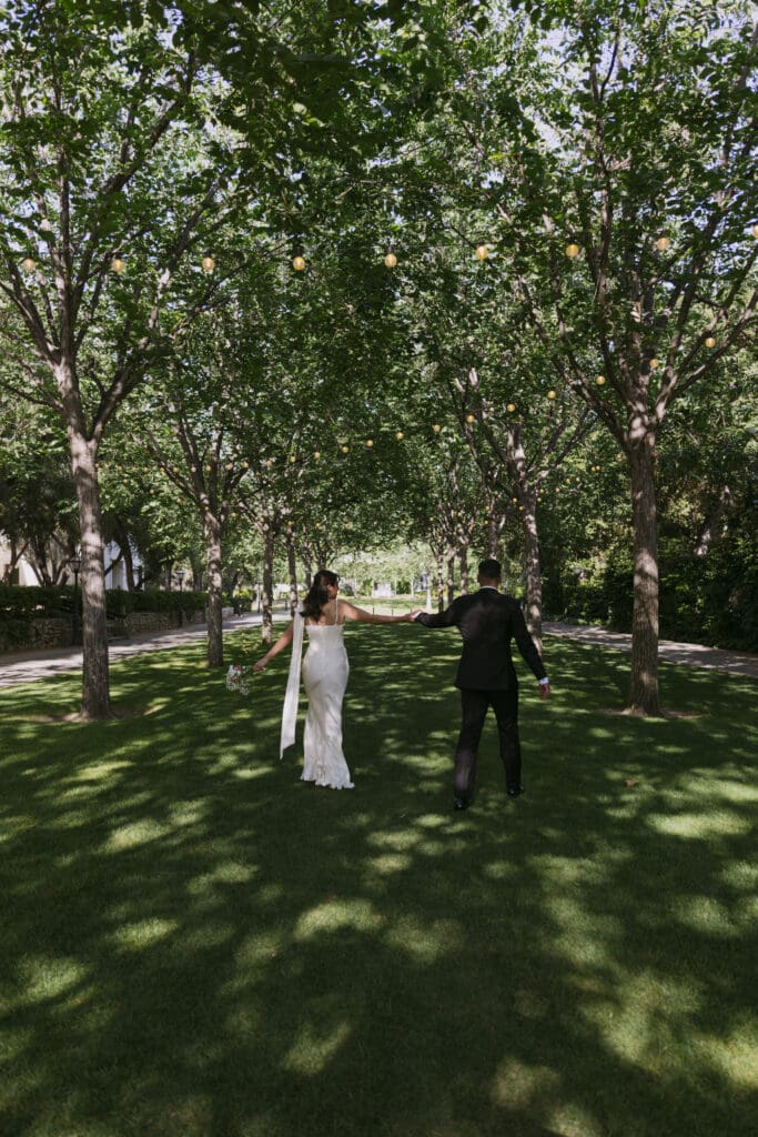 Bride and groom walking under shade from forest of trees.