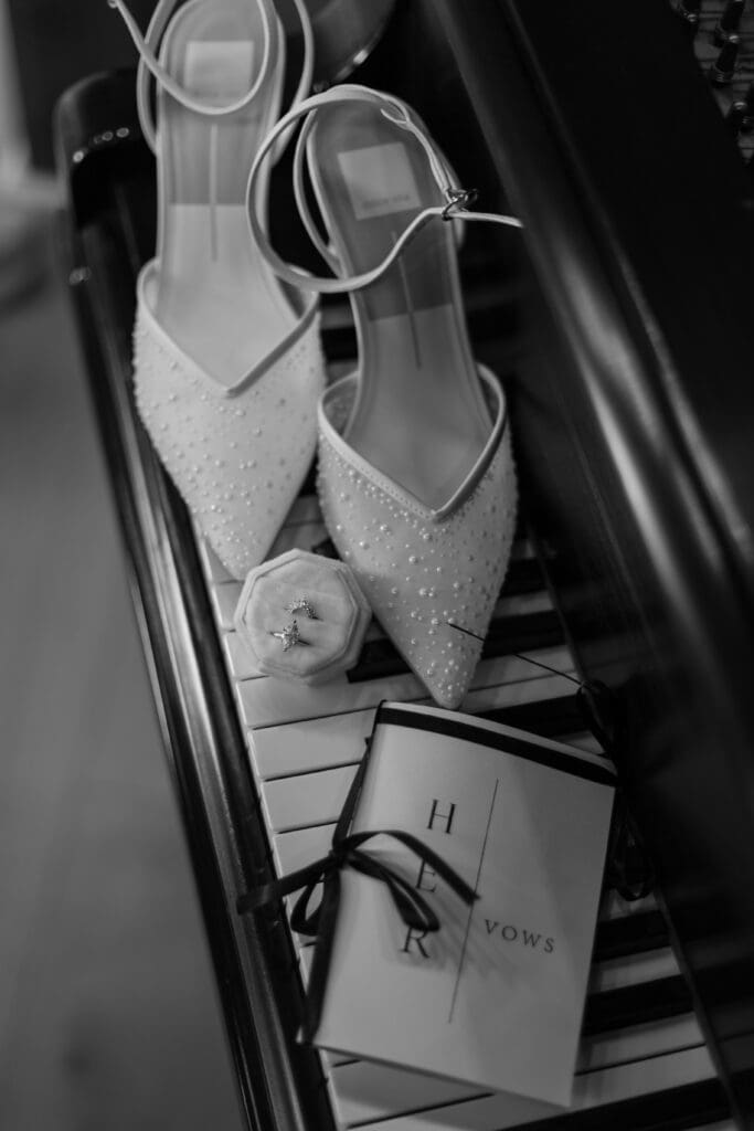 Wedding flat flay on a piano with wedding shoes, rings, and vow book, in black and white.